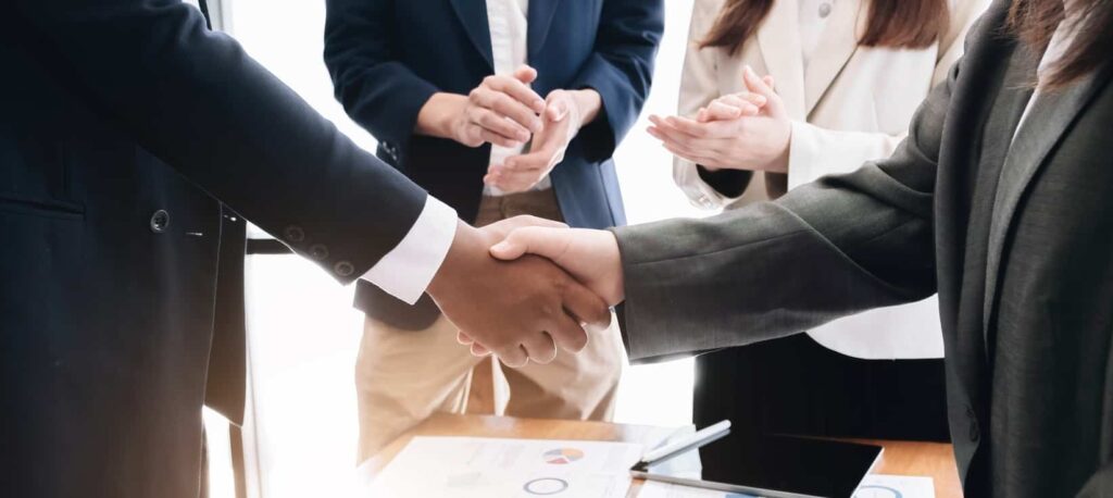 Two business men shaking hands while their colleagues applauding in the background